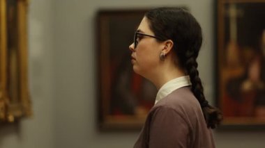 Student young beautiful Caucasian woman contemplating masterpice art. History and culture. Visiting excursion in gallery. Side view. Concept of historic education.