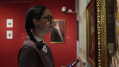 History and education. Side view of pretty young caucasian woman contemplating masterpiece and holding smartphone. Visiting excursion in art gallery. Concept of cultural tourism.
