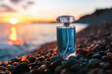 A blue perfume bottle on a pebble beach, against the sunset. Fashionable luxury perfume. Presentation of summer toilet water. The concept of selective perfumery.