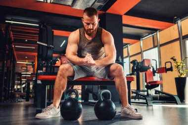 Genç sakallı atletik bir adam spor salonunda dambıllarıyla antrenman yapıyor. Sporcu ellerine magnezya sürer. Zindelik kavramı.