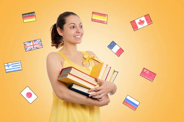 World Book Day. A young smiling female student in a yellow dress holds a stack of books. Yellow background with flags of different countries. The concept of learning foreign languages.
