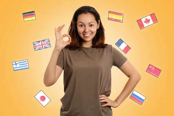 英語の日 若い笑顔の女がOkのジェスチャー 異なる国の国旗を持つ黄色の背景 外国語を学ぶという概念 — ストック写真