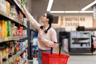 Side view of young Caucasian female student holds basket and reach hand to takes out grocerie from top shelf. Shopping in supermarket and grocery store.