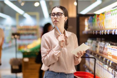 Gözlük takan genç, düşünceli beyaz kadın portresi ürün listesini tutuyor ve etrafa bakıyor. Süpermarkette alışveriş ve tüketim kavramı.