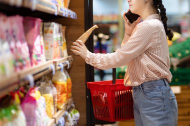 Shopping. Woman holds product and calls on phone. Girl talking on smartphone choosing food in supermarket. Side view.