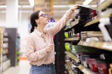 Side view of young Caucasian woman wearing glasses take baking dish from upper shelf. Shopping and choice of kitchen tools.