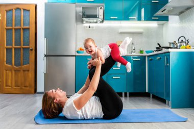 Gülümseyen bir anne, minderde spor yaparken sevimli mutlu bir bebeği kollarında tutar. Uzayı kopyala Çocuklarla evde spor eğitimi kavramı.