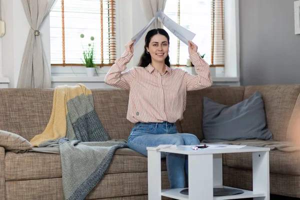 stock image Smiling pretty young Caucasian woman sitting on sofa and holds documents over her head in form of roof. Concept of loan and mortgage.