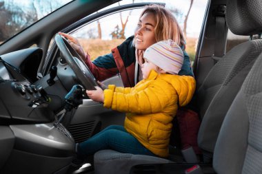 A young smiling mother teaches her little child to drive a car. The kid is sitting in a chair and holding the steering wheel with his hands. Side view.