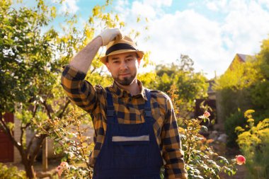 Portrait of a bearded young smiling gardener wearing a straw hat. In the background there is a backyard and a garden. The concept of gardening and horticulture.