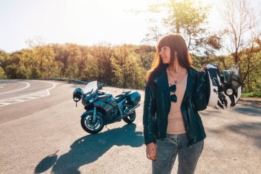 Deri ceketli, elinde kaskla gülümseyen genç bir kadın. Arka planda, bir motosiklet ve bir yol. Motosiklet yerel seyahat kavramı.