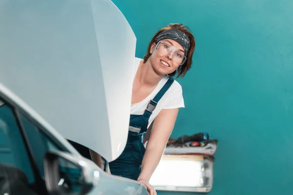 Portret Młodej Uśmiechniętej Mechaniczki Okularach Kombinezonach Wychodzącej Zza Maski Samochodu — Zdjęcie stockowe
