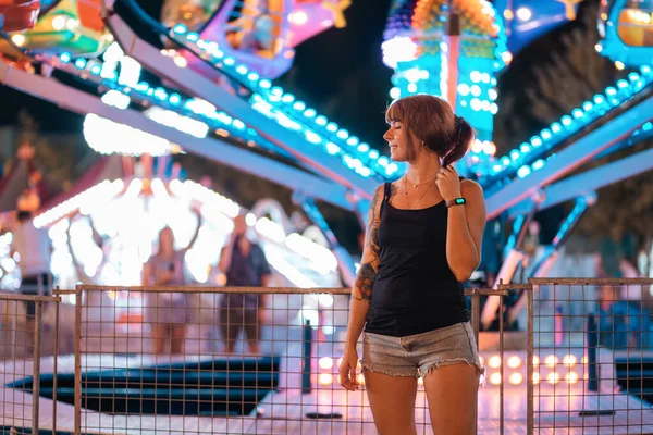 Holiday. A young happy woman with tattoos is resting in an amusement park. In the background there is a carousel glowing with neon lights.