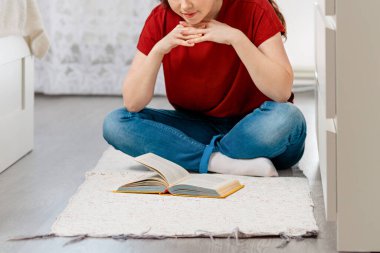 A young Caucasian woman sits cross-legged on the floor and reads a book with interest. Close up. In the background, the white interior of the room. The concept of education and reading books.