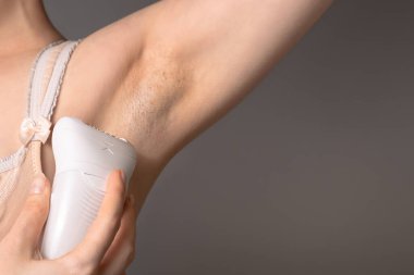 A young woman removes excess hair with a depilator in the armpit area. Concept of home care and Spa. Copy space and close up. Gray background.