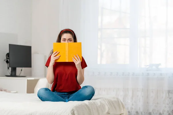 Kobieta Siedzi Łóżku Zakrywa Twarz Żółtą Książką Jasny Pokój Komputer — Zdjęcie stockowe