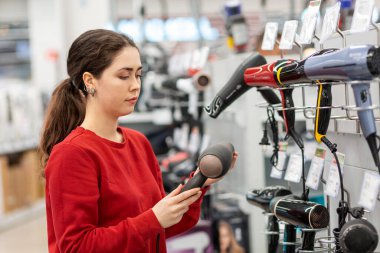 Caucasian young woman with long hair, holding a hair dryer. In the background, shelves with household appliances. The concept of buying household appliances for beauty and self-care.
