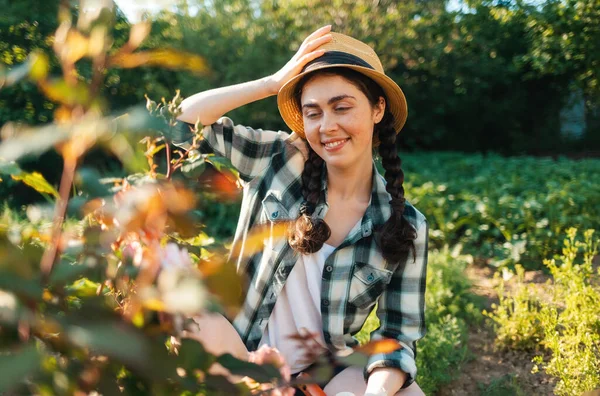 Portrait of smiling young woman in a straw hat sits in the garden, looking at flowers of a rose.