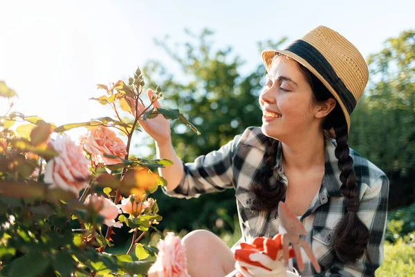 戴着草帽的年轻快乐女子的画像坐在花园里 触摸着玫瑰的花朵 夏季园艺 — 图库照片