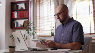 Caucasian bald bearded young man is counting American dollars in cash. Manager sitting at desk with laptop. Concept of accounting and tax refund.