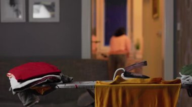 Close up of stack of clothes and steaming hot iron left on ironing board. In background defocused housewife doing chores. Home chores.