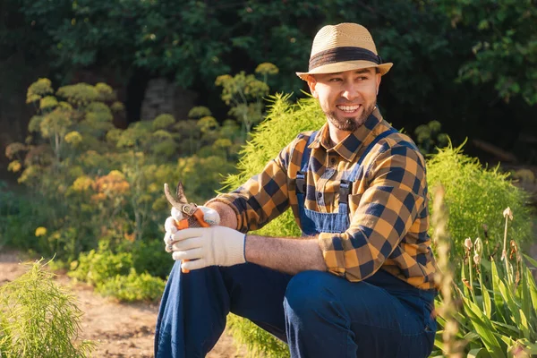 Portrait of bearded, handsome happy gardener in uniform and a straw hat sits and relax at his kitchen-garden, holding a pruner. In the background is a backyard and a garden. The concept of gardening and spring.