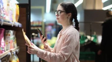 Genç beyaz kadın portresi süpermarkette ürünleri seçiyor ve QR kodlarını tarıyor. Modern alışveriş yemekleri.