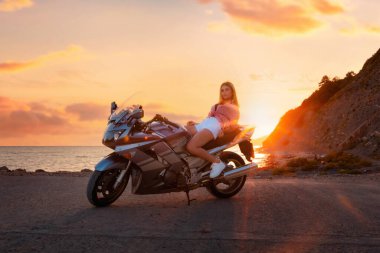Dünya Motosiklet Günü. Beyaz, genç bir kadın motosiklet üzerinde poz veriyor. Arka planda altın gün batımı ve okyanus. Özgürlük ve motosiklet gezileri.