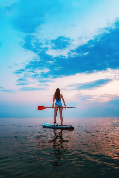 一个年轻的 晒黑了的 纤细的女人站在一块锯齿状的木板上 船桨在海中 夕阳西下垂直的 冲浪的概念 — 图库照片