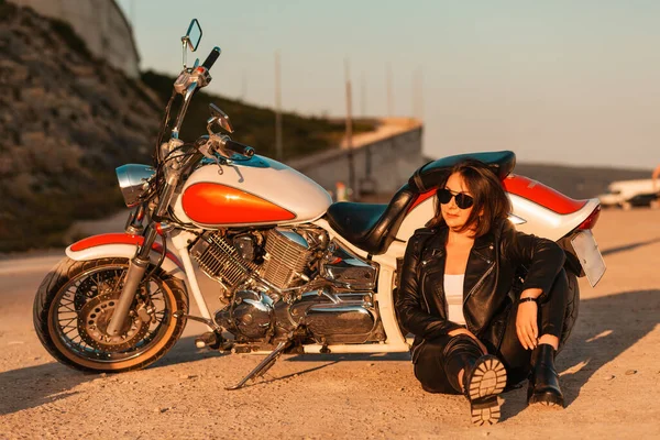 Cool confident woman in a leather motorcycle outfit sitting on the ground near a motorcycle. Sunlight. The concept of the Motorcyclist Day.
