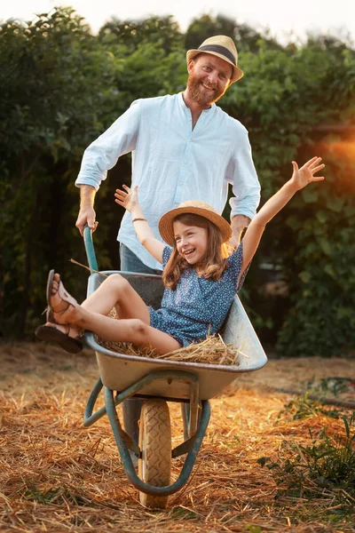 stock image Happy bearded father in a straw hat is pushing a cart with a cheerful little daughter. Family weekends together in the backyard. The concept of gardening.