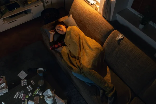 stock image Flu and fever. Top view of illness Caucasian woman lies on sofa covered with blanket. Table with medicine and pills. Loneliness and depression concept.