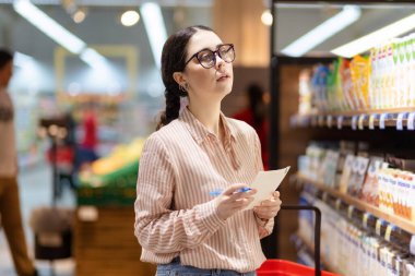 Gözlük takan genç beyaz kadın ürün listesi tutuyor ve market çeşitlerinden birini seçiyor. Süpermarkette alışveriş ve tüketim kavramı.