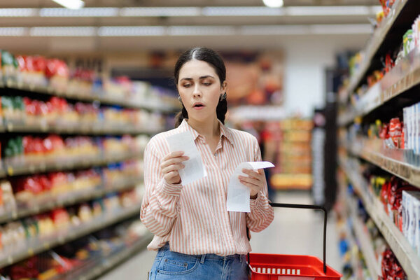 Portrait of young amazement Caucasian woman by high prices in supermarket. Shocked millennial holds receipts of purchase. In background, rows of shelves with products. Concept of inflation.
