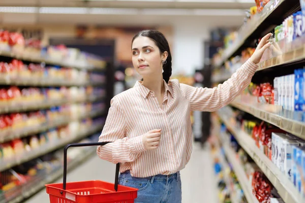 年轻迷人的高加索女人的画像手握购物车 从架子上取食 在背景上展示 超市购物的概念与消费主义 — 图库照片