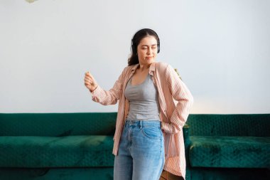 Oturma odasında dans eden, kulaklık takan beyaz bir kadının portresi. Arka planda yeşil kanepe. Ev eğlencesi.