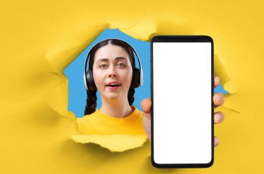 Sarı kağıt arka planda yırtılmış bir delik, kulaklık takan mutlu genç kadın beyaz ekranlı akıllı telefonun yakın çekimini gösteriyor. Model yap ve uzayı kopyala. Teknik destek, çevrimiçi öğrenim ve eğitim kavramı.