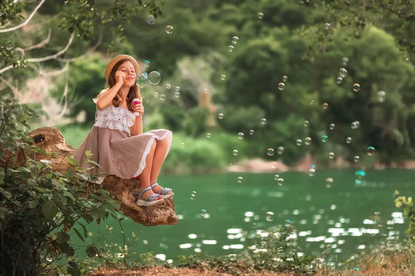 暑假The Summer Time Holidays 可爱的小女孩坐在原木上吹着肥皂泡 气泡四处飞扬 复制空间 快乐童年及儿童节的概念 — 图库照片