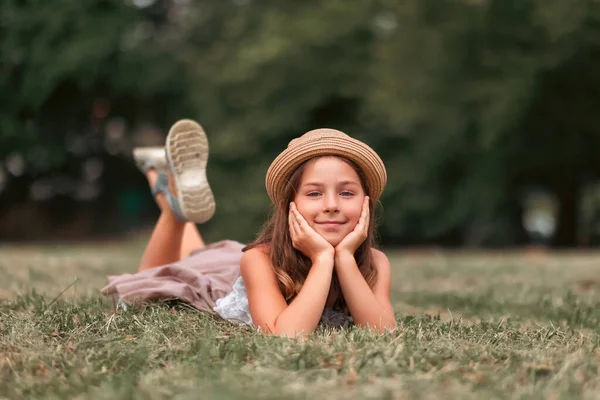 stock image Portrait of cute smiling girl in a straw hat lies on the lawn and looks around. Copy space. The concept of school holidays and summertime.