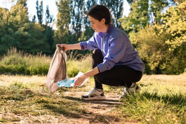 Spor kıyafetli genç bir kadın plastik şişeleri alıp çöp torbasına koyuyor. Yan görüş. Çevre koruma ve gönüllülük kavramı.