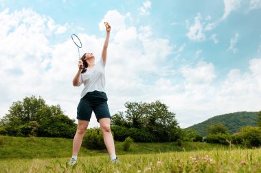 Genç, sportif bir kadın elinde raket tutuyor ve bir mekik horozuna vurmaya hazırlanıyor. Uzayı kopyala Arka tarafa park et. Badminton oyunu ve boş zaman aktivitesi.