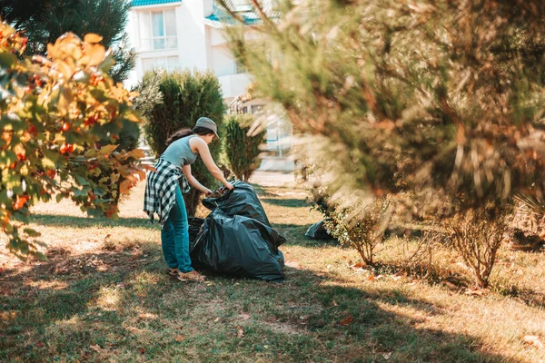 The concept of environmental pollution and Earth day. Woman volunteer doing cleaning in the Park in front of the house. She\'s putting the garbage bags in one pile. Copy space.
