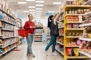 Satış danışmanı ürünü alıcı için üst raftan alır. İki gülümseyen beyaz kadın. Alışveriş, iş ve ticaret kavramı.