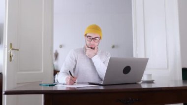 Gözlük takan genç, sakallı, beyaz bir adam belgeye yazılıyor ve dizüstü bilgisayarda çalışıyor. Uzaktan çalışma, serbest çalışma ve çevrimiçi halkla ilişkiler kavramı