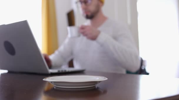 手前のカップを閉じてください 眼鏡をかけた髭の男がノートパソコンで働いてコーヒーを飲んでいる リモートワーク フリーランス オンライン時間の概念 — ストック動画