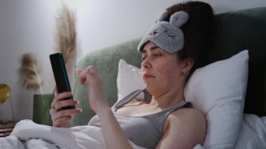 Uyku maskesi takan yorgun beyaz bir kadın rahat bir yatakta uzanıyor ve akıllı telefon kullanıyor.