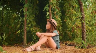 Çocukluk. Hasır şapkalı sevimli bir kız bahçedeki çimlerin üzerinde oturuyor. Yakınlaştır. Arka planda tutkulu meyve tarlaları.