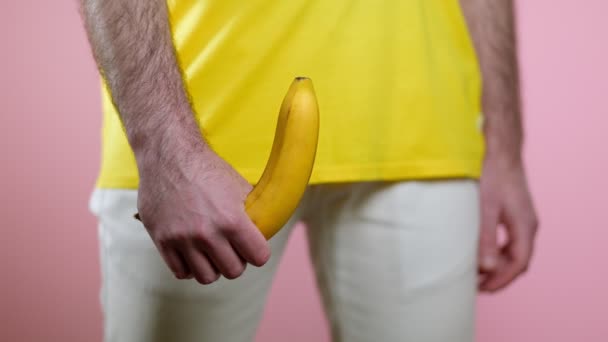 男は性器のレベルで彼の手に上昇したバナナを保持しています 女性の手はバナナの上に指を走らせる ピンクの背景 男性の健康の概念 — ストック動画