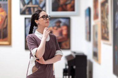 Sergi gezisi. Beyaz tenli, gözlüklü genç bir kadının portresi sanat üzerine kafa yoruyor. Arka planda odaklı olmayan resimler. Eğitim kavramı