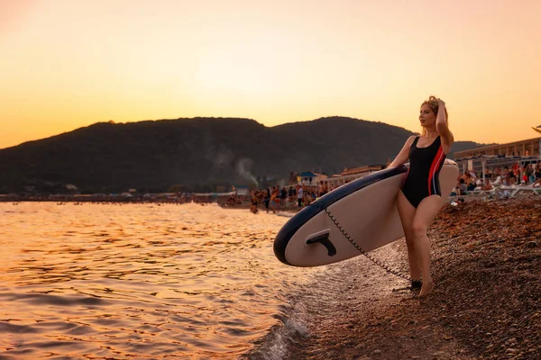 ポーズをとっている若い女性はビーチでボードを持っています コピースペース 背景に沈む夕日 スポーツ 休暇の概念 — ストック写真